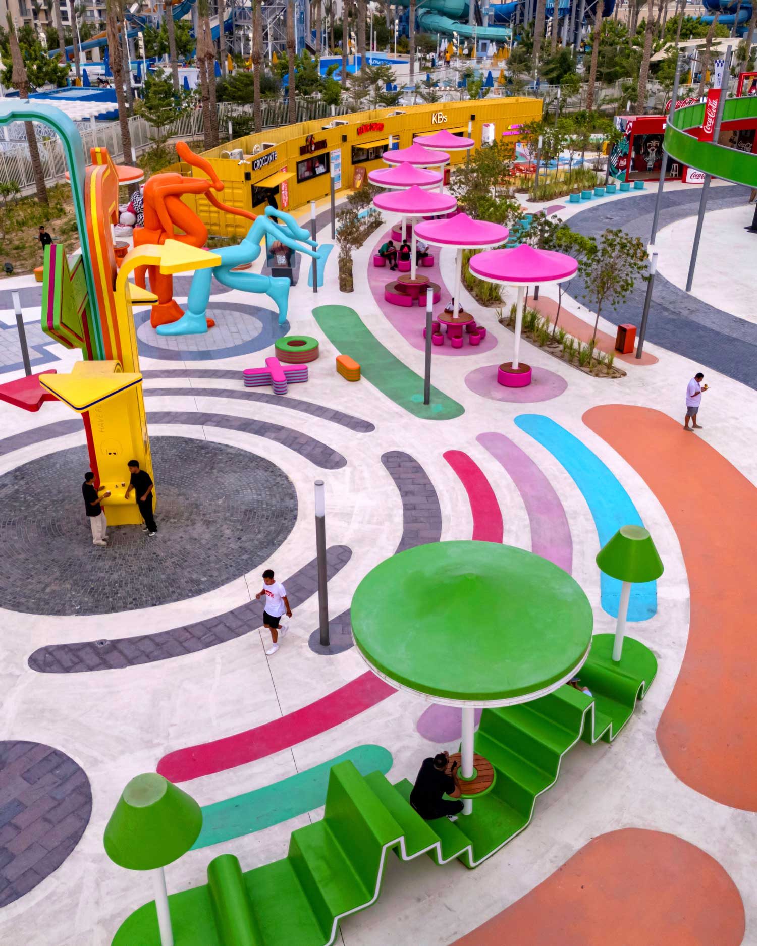 A plaza of shipping-container units and yellow parasols on a swirling coloured pavement.