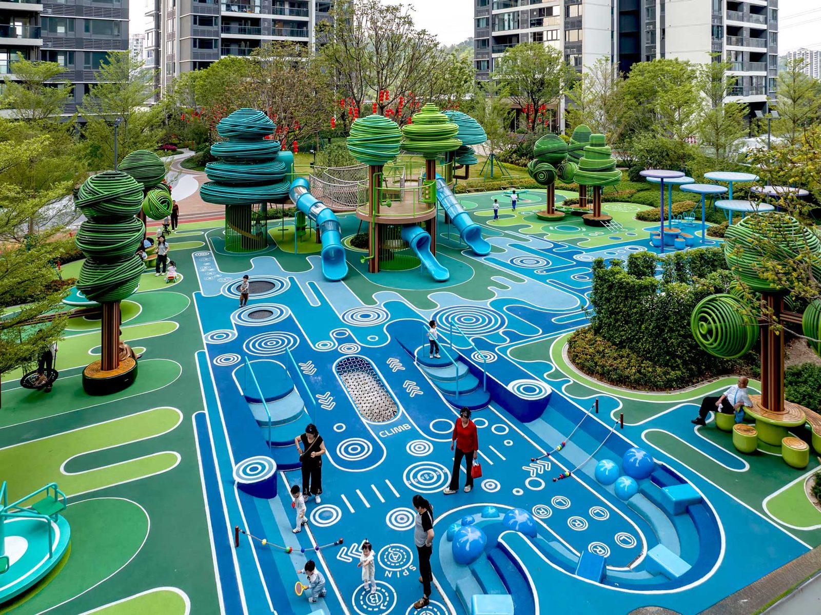 The playscape seen from above with people walking across its blue and green flowing ground pattern.