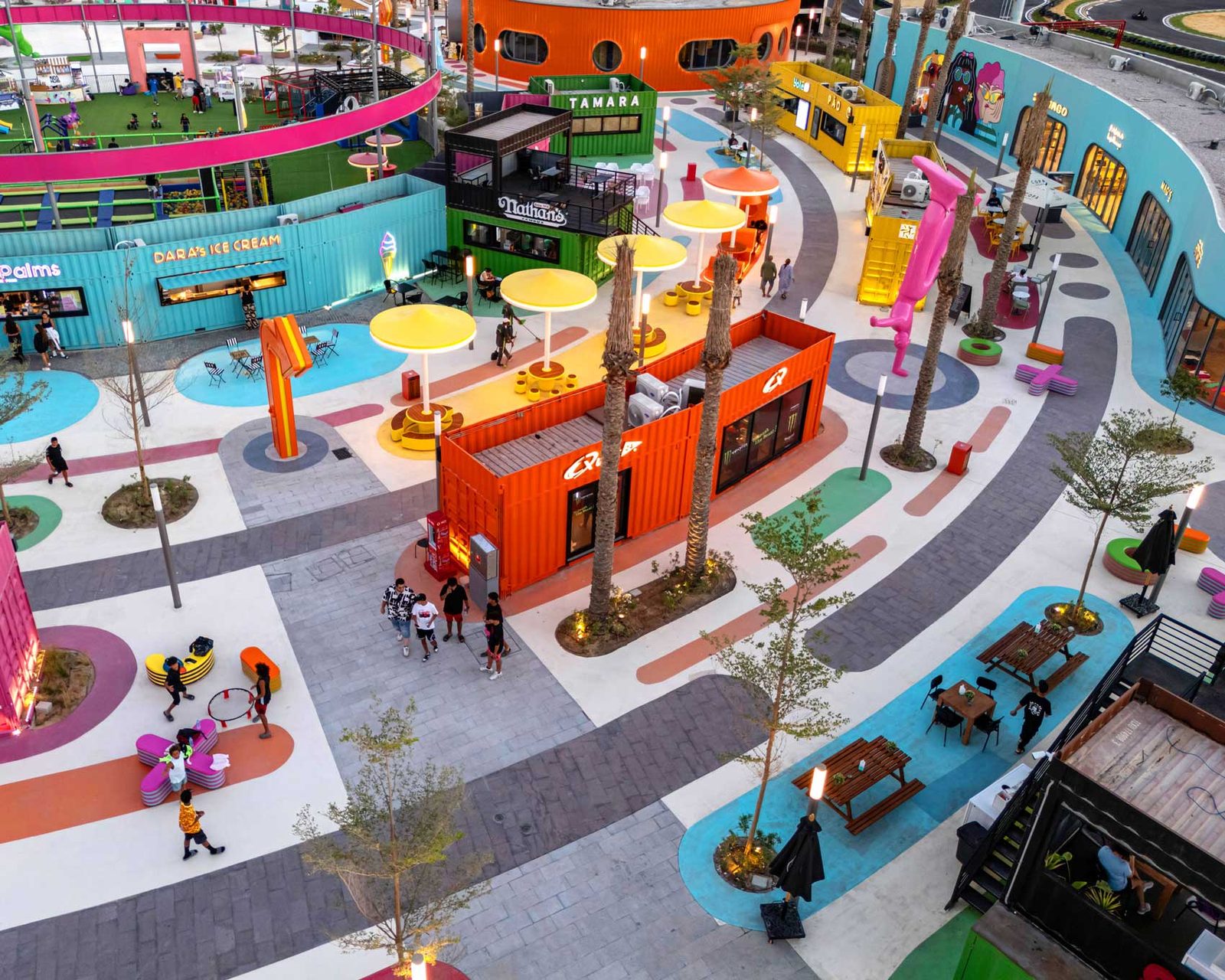 People walking past illuminated container units and coloured canopies on the swirling pavement at dusk.