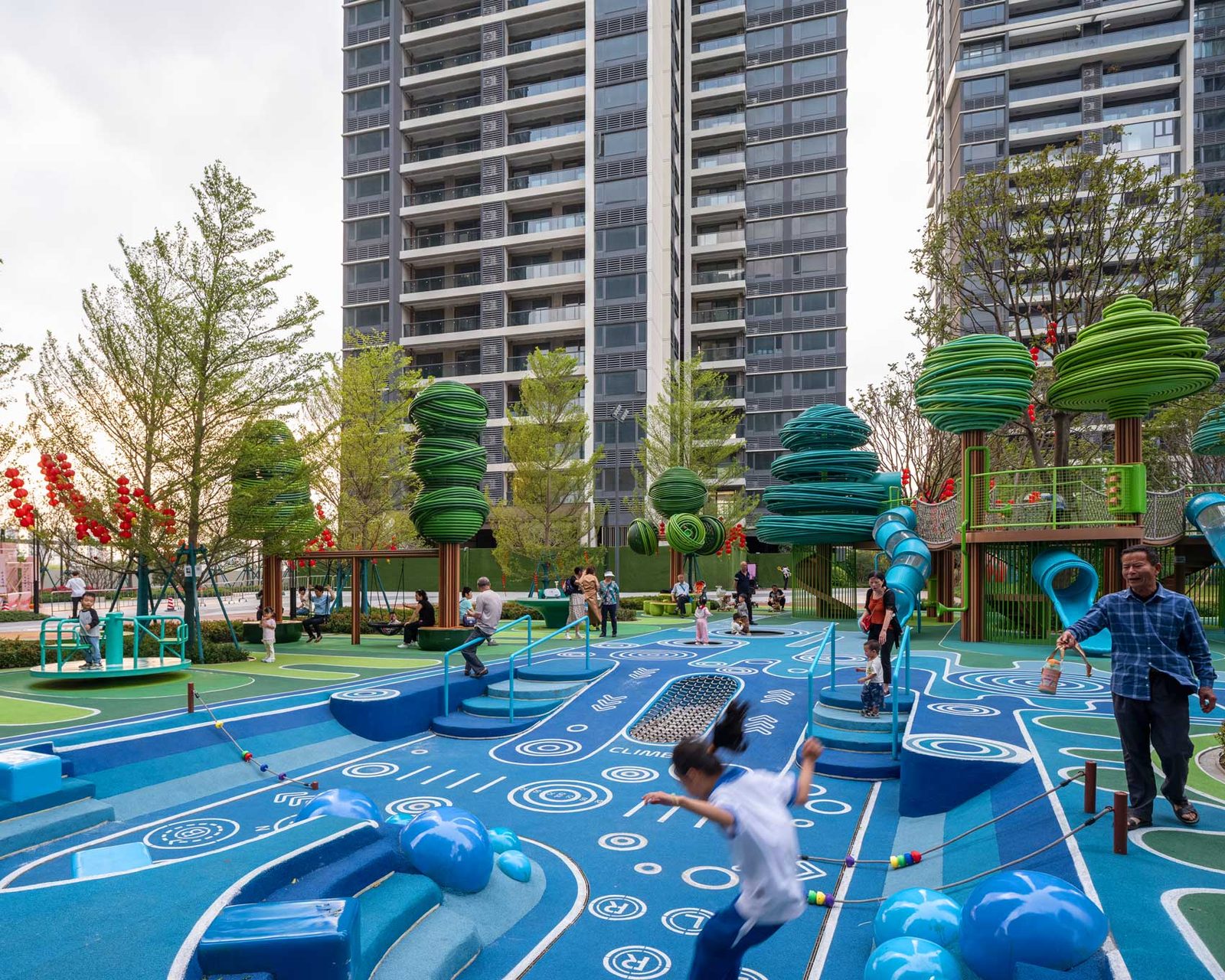Children and adults moving across the blue and green flowing ground surface beneath the sculptural trees.