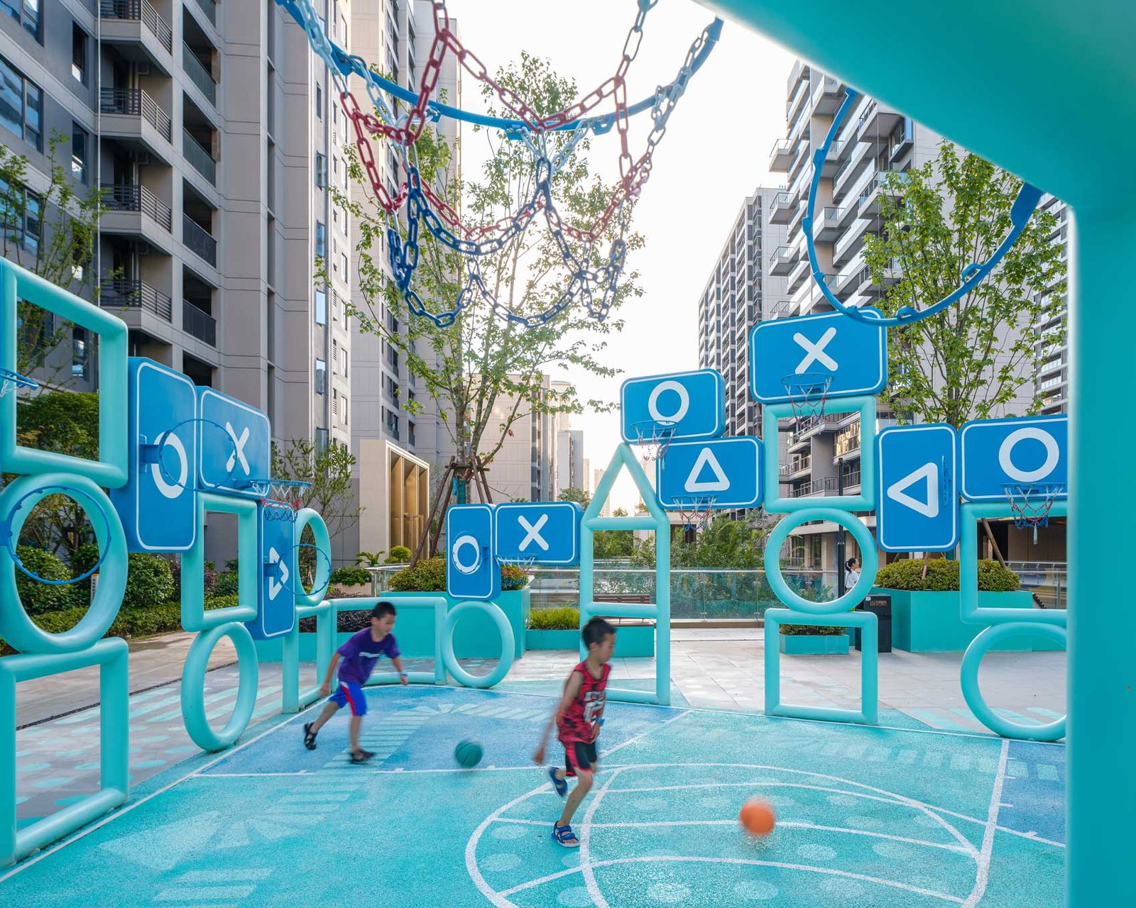 Two children running after a ball inside a turquoise court framed by panels printed with circles, crosses and triangles.