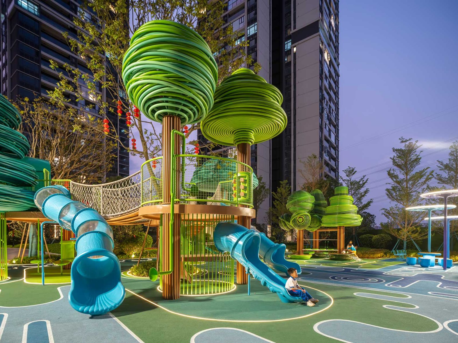 Two sculptural green tree structures with slides and a hanging bridge, photographed at dusk above the green and blue play surface.