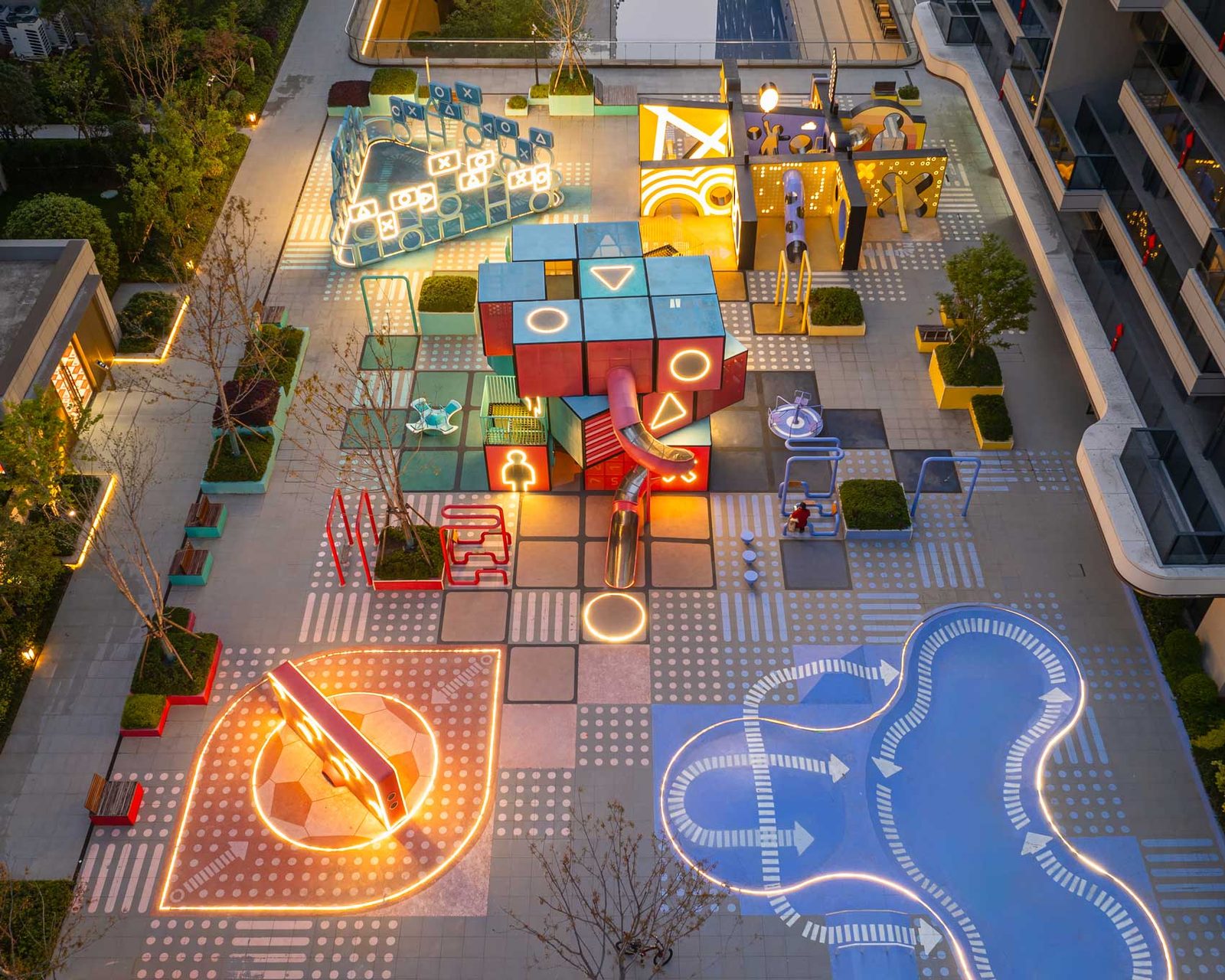 The plaza at dusk, its illuminated ground graphics and coloured cube volumes running between residential towers.