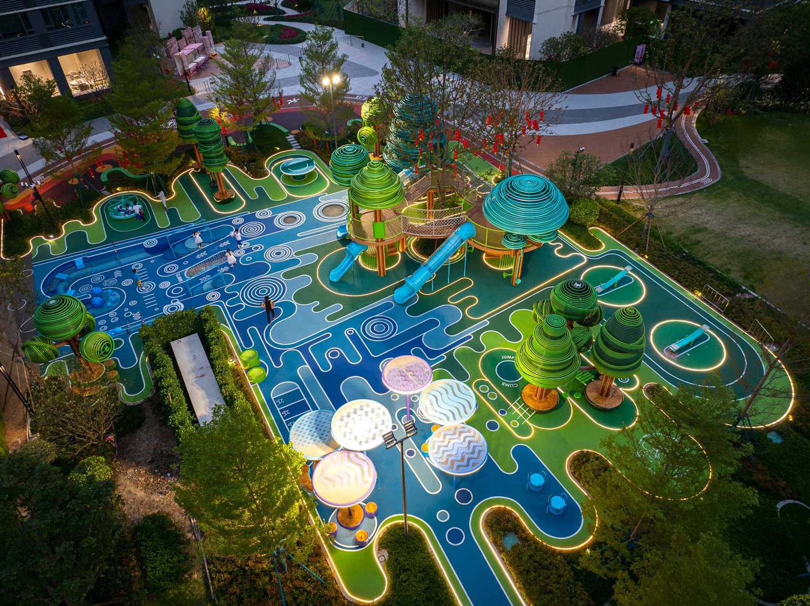 Evening aerial view of the illuminated play surface set within planting and residential blocks.