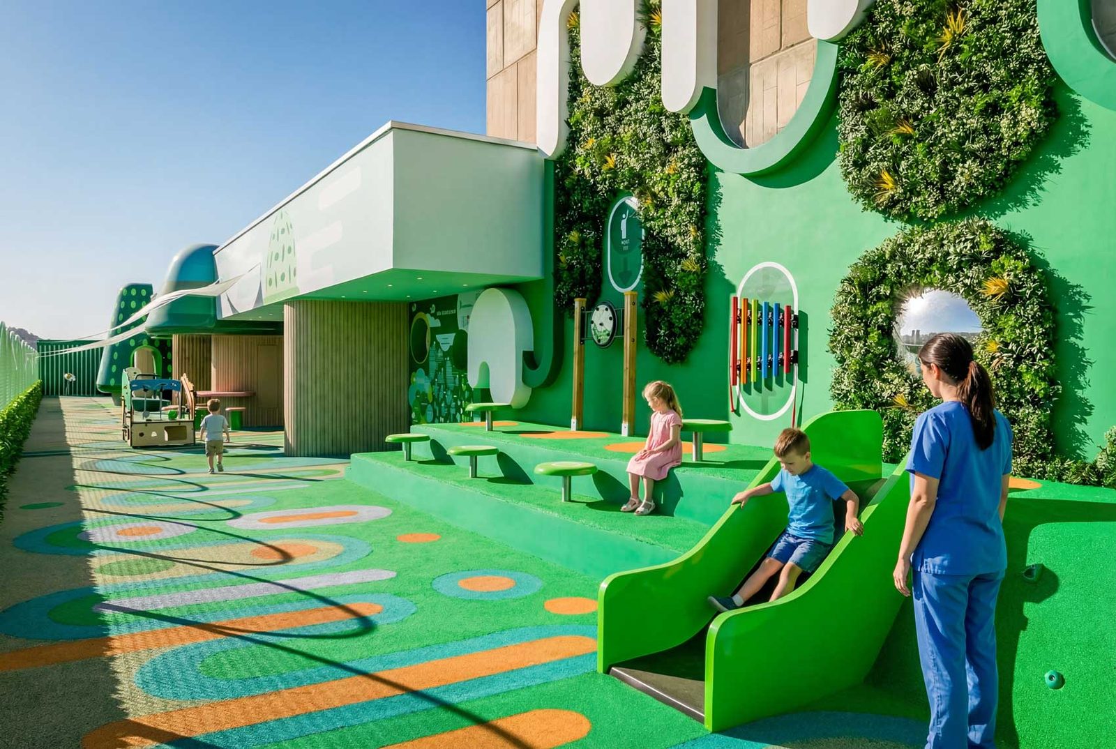 Children and a member of hospital staff on the green terrace, with rounded green canopies and a slide.